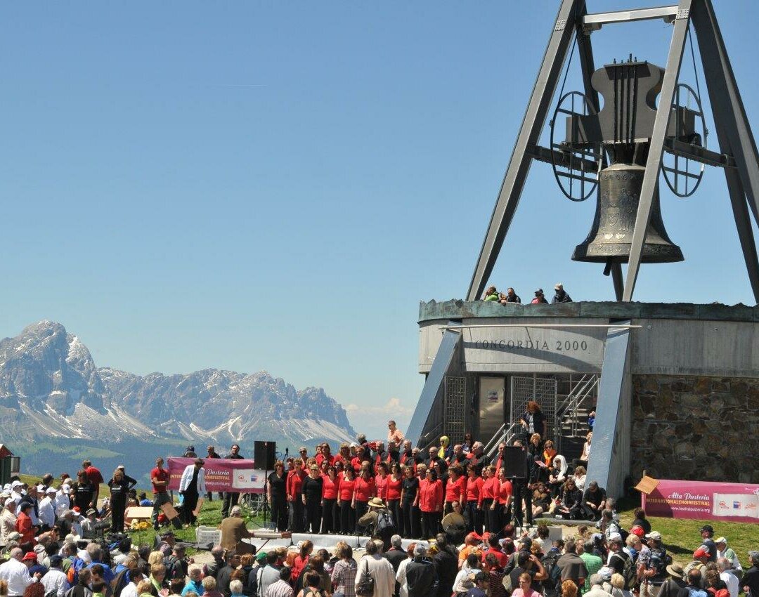 Ein Chor singt vor Publikum vor der Glocke Concordia 2000 am Kronplatz-Gipfel mit dem Peitlerkofel im Hintergrund. | © Skirama Kronplatz