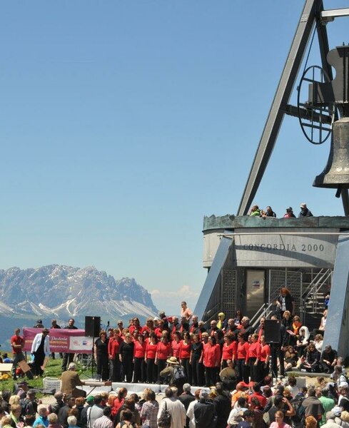 Ein Chor singt vor Publikum vor der Glocke Concordia 2000 am Kronplatz-Gipfel mit dem Peitlerkofel im Hintergrund. | © Skirama Kronplatz