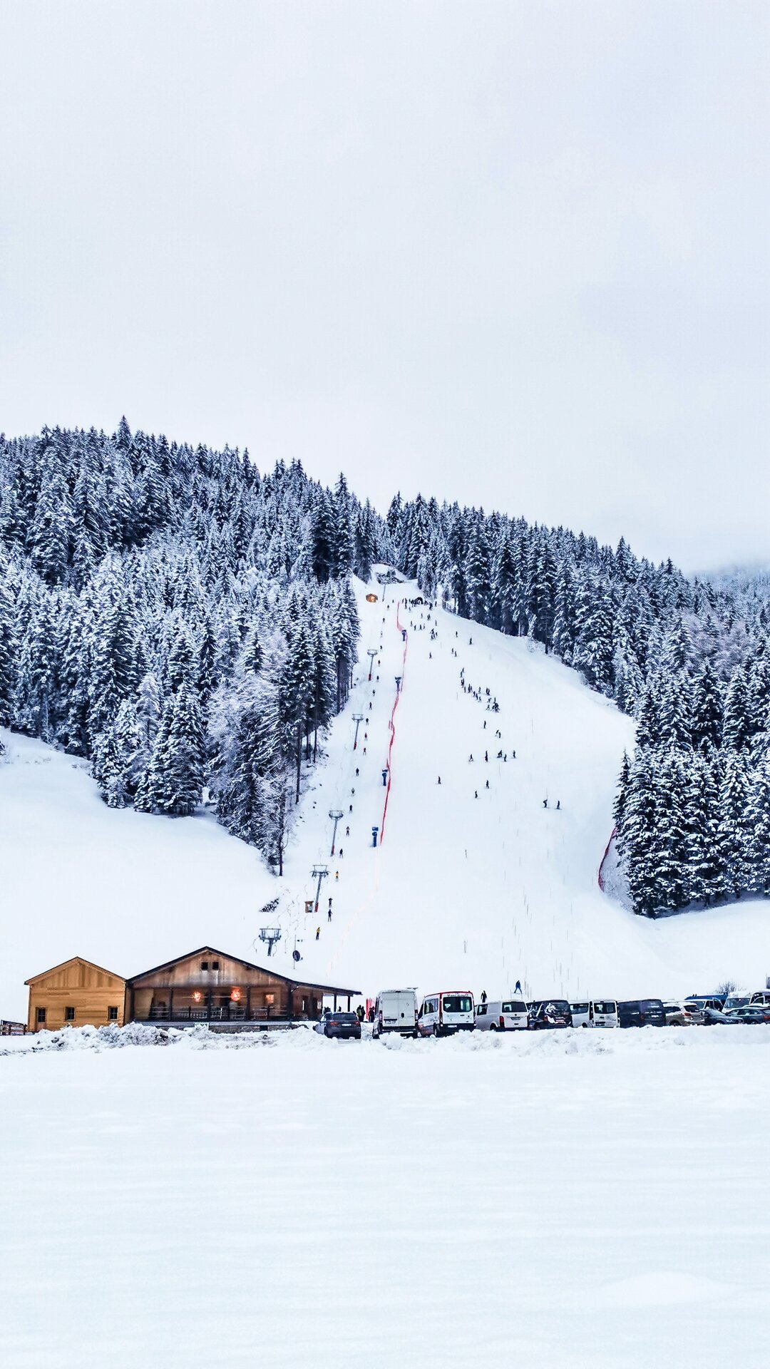 Ein Holzhaus mit einem neuen Anbau aus Holz, rechts daneben einige PKWs, steht am Fuße eines verschneiten, bewaldeten Berges mit einigen Skifahrern auf der Piste und mit bewölktem Himmel im Hintergrund. | © Skilift Pichl