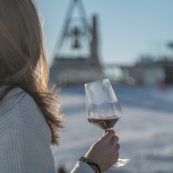 Eine Frau schaut auf die Concordia Glocke am Kronplatz Gipfel mit einem Glas Rotwein in der Hand. | © Gianvito Coco