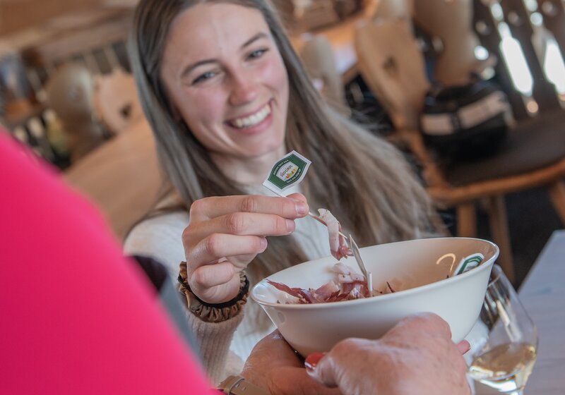Eine Frau nimmt sich ein Stückchen aufgeschnittenen Speck mit dem Zahnstocher aus einer kleinen Schüssel, die ihr eine andere Person hinhällt. | © Gianvito Coco
