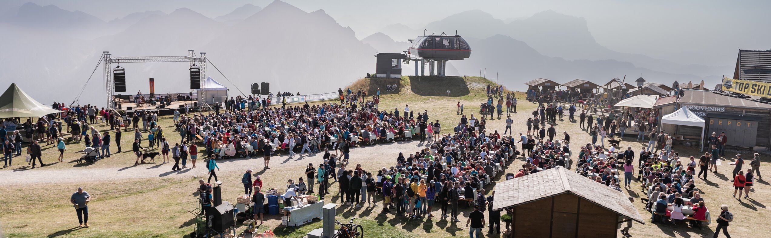 Zahlreiche Menschen versammeln sich auf einer Wiese vor Bühne und Marktständen mit Bergkulisse im Hintergrund. | © Gianvito Coco