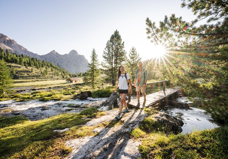 Zwei Wanderer gehen über einen von Sonnenstrahlen durchfluteten, felsig-grünen Pfad in alpiner Landschaft. | © Harald Wisthaler