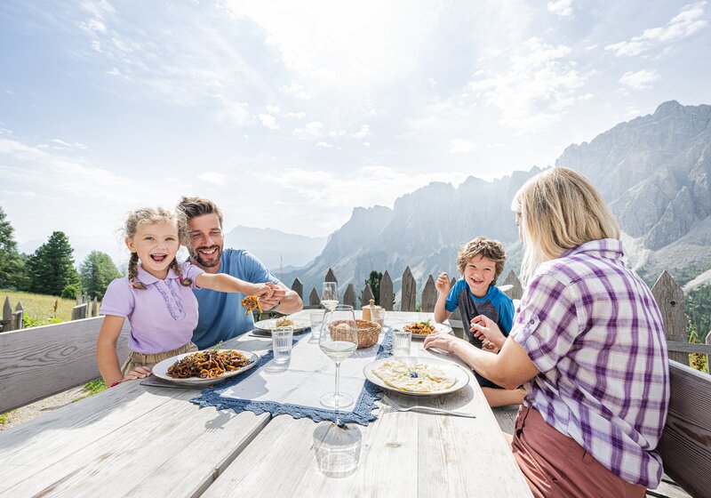 Eine glückliche Familie genießt eine Brotzeit auf einem Holztisch unter freiem Himmel, im Hintergrund die Berge, Sonne und einzelne Wolken. | © Harald Wisthaler